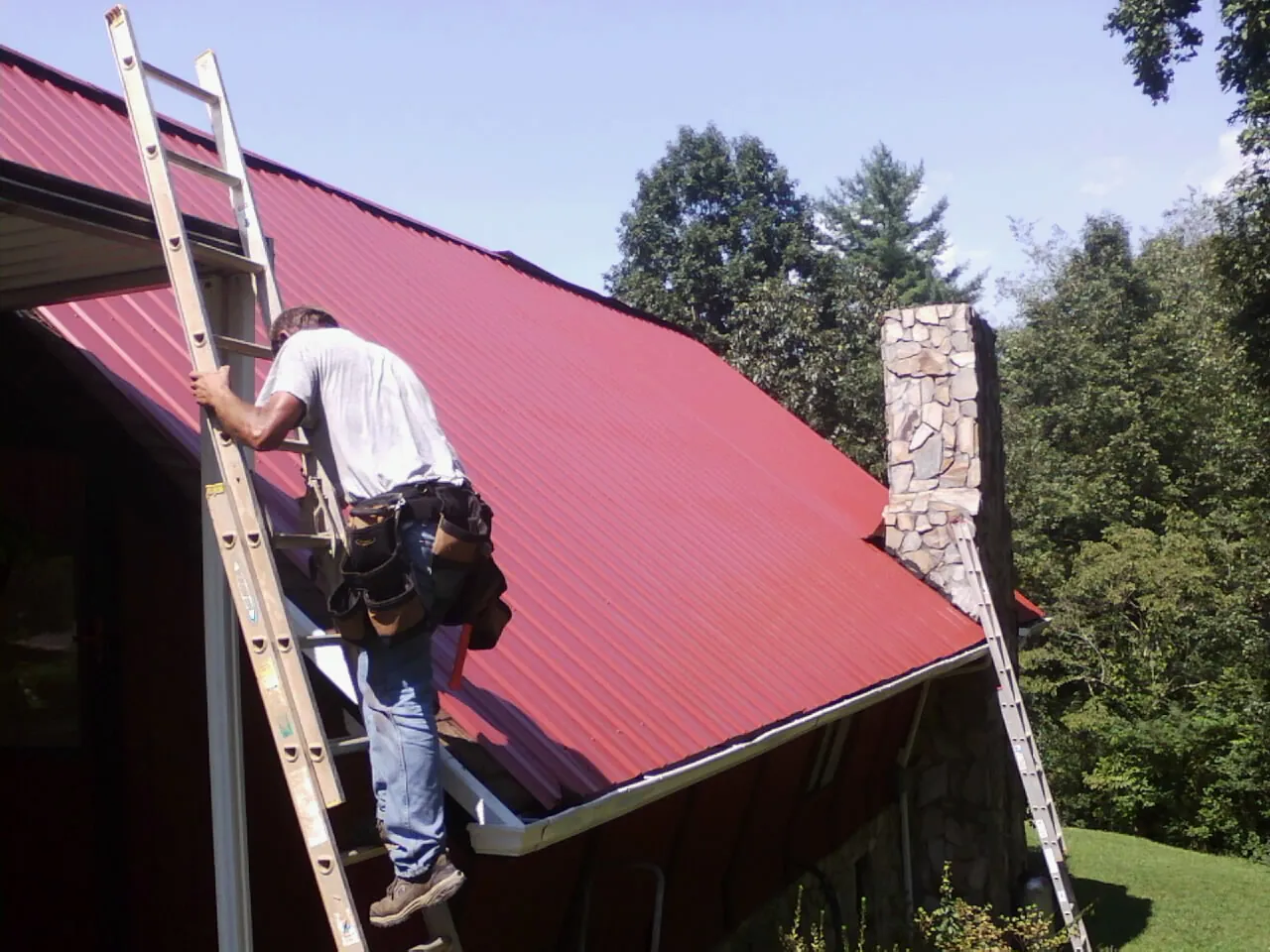 Roofer on a ladder beside a red metal roof and stone chimney — Soffit & Fascia Repair by Roofing Brothers in Piedmont